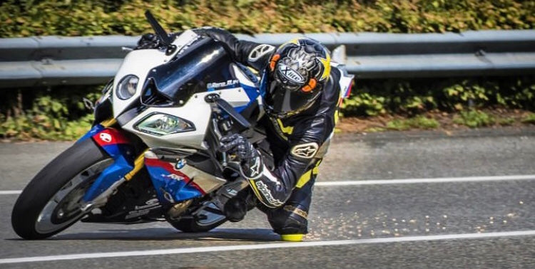 Motorradfahrer auf einem weißem Motorrad in einer Linkskurve. Er trägt ein Dymostar Knieschleifer am Knie und schleift damit auf der Straße. Es sind sehr viele helle Funken hinter Ihm zu sehen.