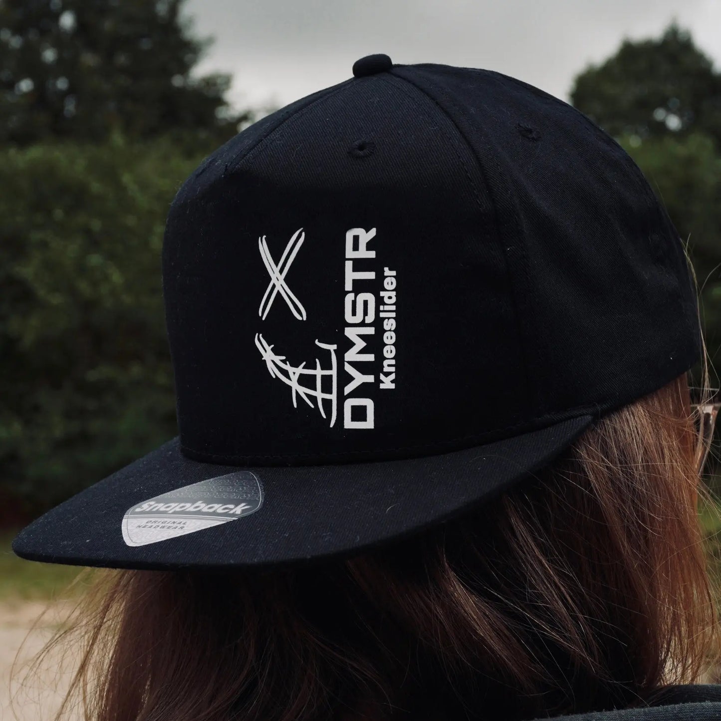 Schwarze DYMSTR SnapBack für Motorradfahrer mit weißem Firmenlogo „DYMSTR / DymoStar“ auf der Vorderseite.